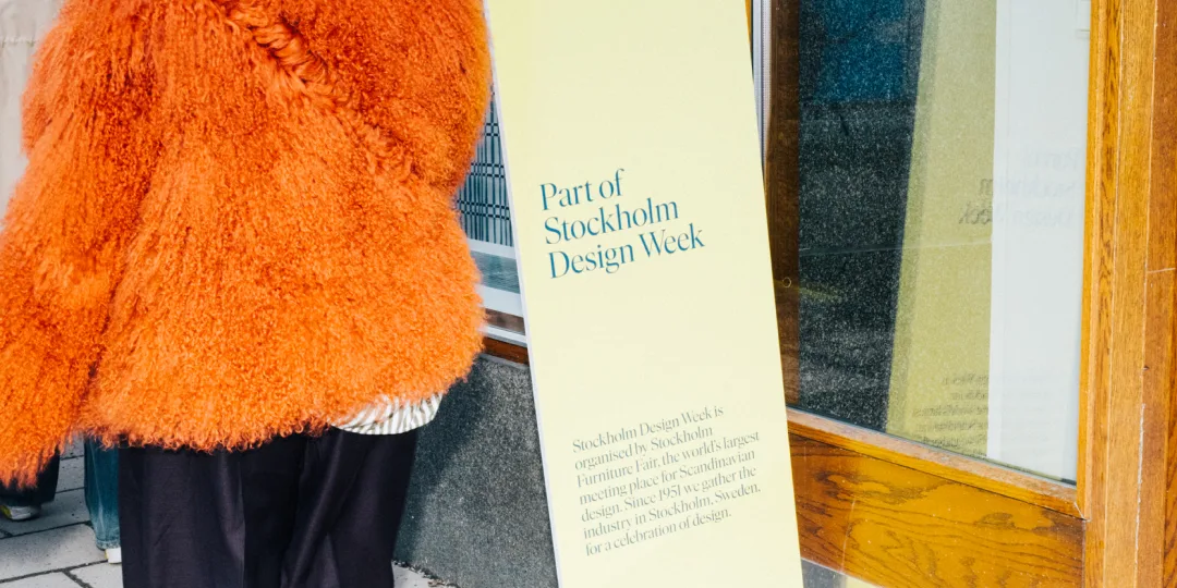 A person in an orange coat standing outside a showroom featuring a sign for Stockholm Design Week participation.